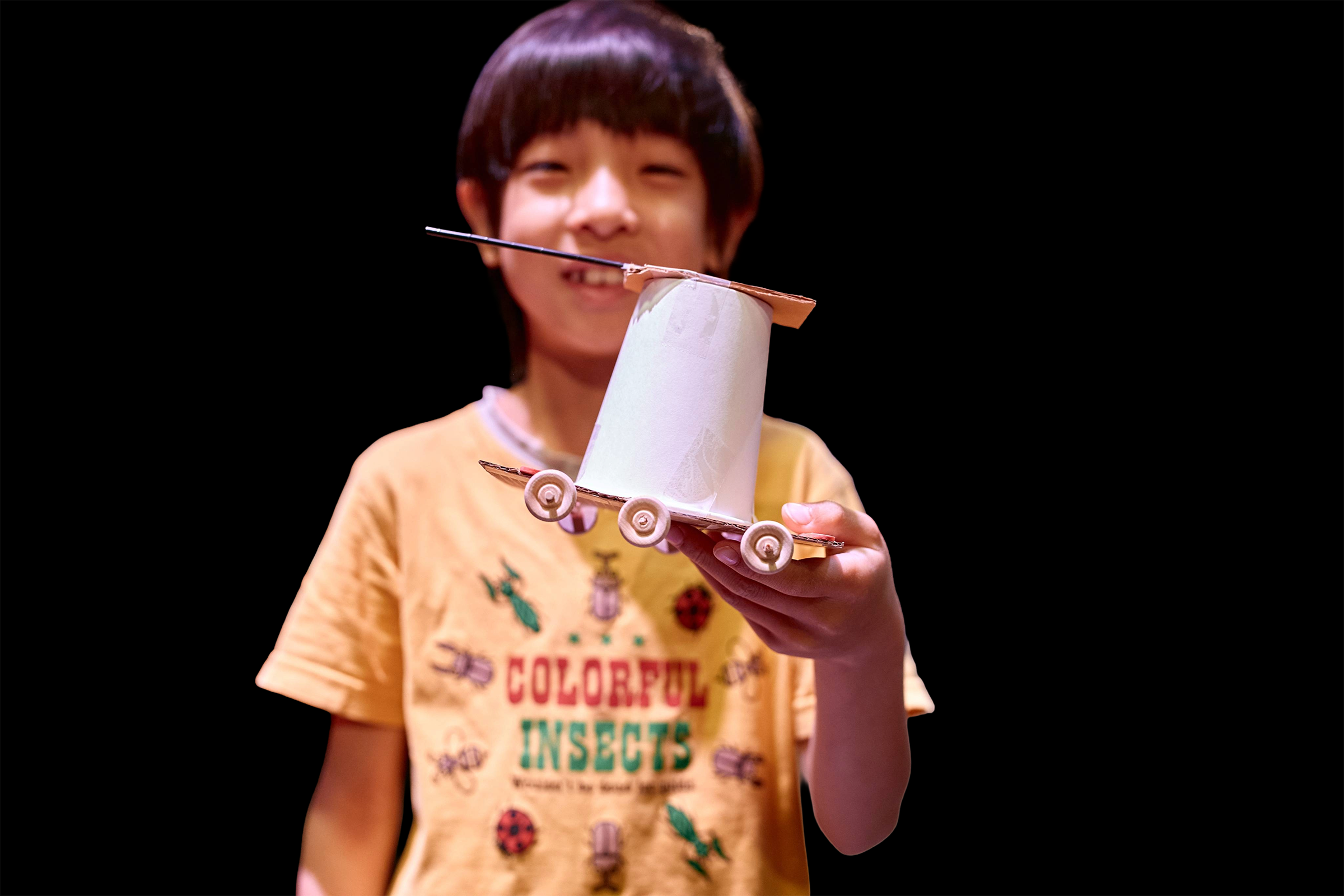 Smiling boy in a yellow t-shirt showing off his arts and crafts project - a car made of a paper cup and wooden wheels